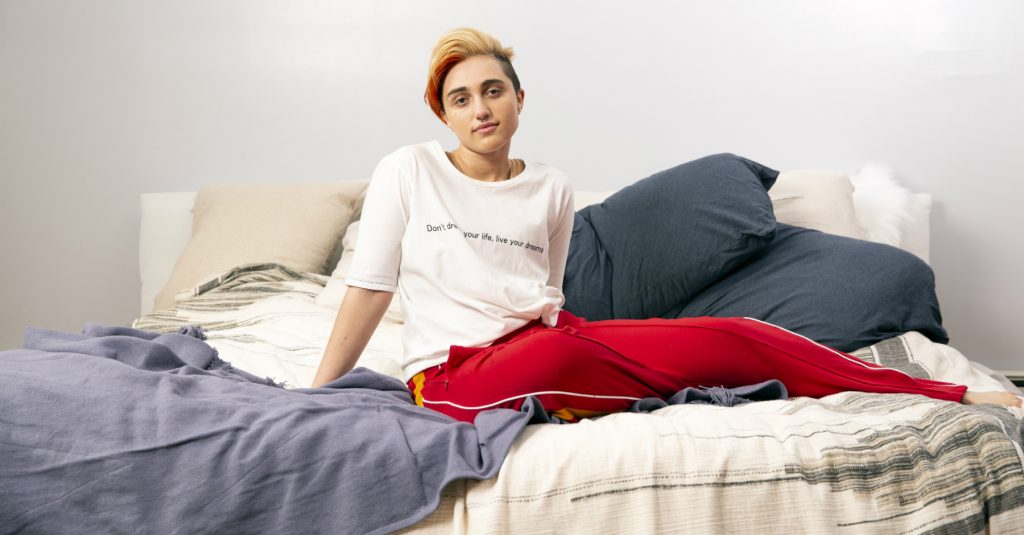 A young person sitting on a bed, looking calmly toward the camera