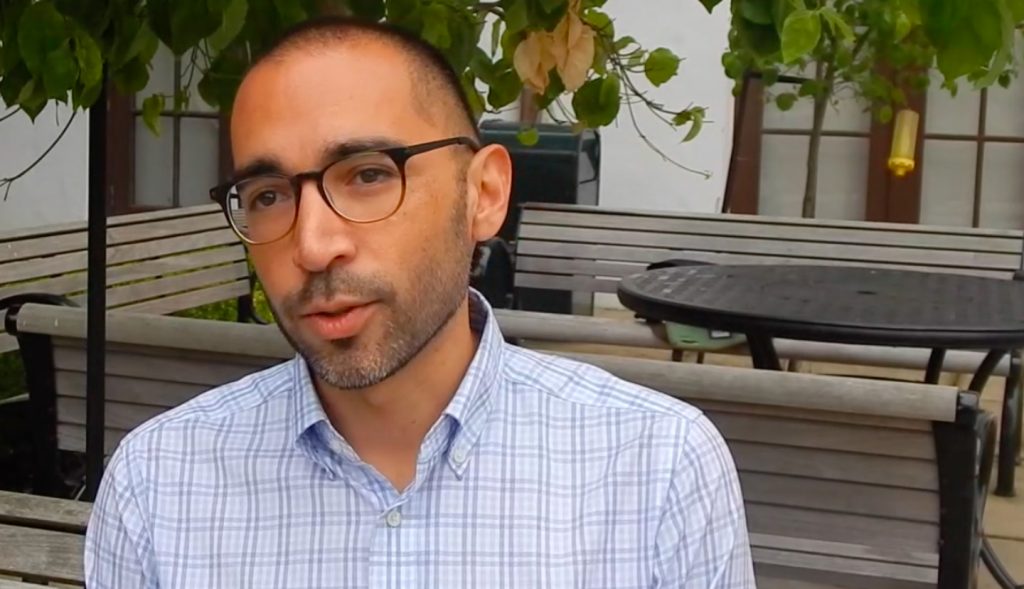A man speaking during a filmed interview on an outdoor terrace